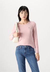 Ljusrosa ribbad långärmad T-shirt med rund halsringning, tillsammans med blå denimjeans. Personen håller en krämfärgad väska.