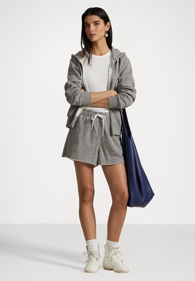 Grijze ritsloze hoodie over een witte geribbelde top, gecombineerd met grijze shorts. Accessoires omvatten een grote blauwe tas en witte high-top sneakers.
