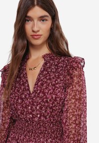 Robe florale bordeaux avec des manches longues transparentes, des épaules volantes et une taille smockée. Présente de petits motifs floraux roses et blancs.