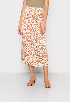 Kobieta w beżowym topie na ramiączkach, białej spódnicy do połowy łydki z pomarańczowym abstrakcyjnym nadrukiem zwierzęcym oraz brązowych sandałach na obcasie z odkrytymi palcami, stojąca na szarej podłodze.