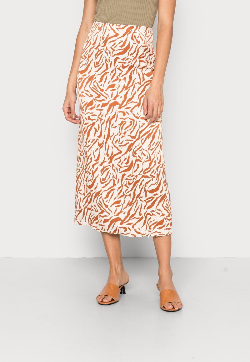 Femme portant un débardeur beige, une jupe midi blanche avec un imprimé animalier abstrait orange, et des sandales à talons ouvertes couleur beige, debout sur un sol gris.