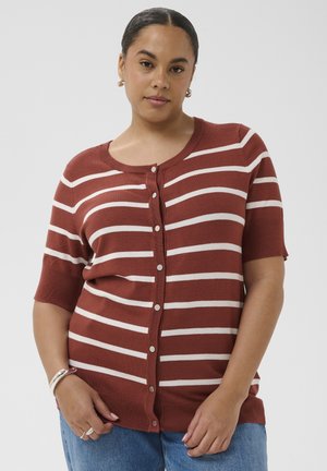 Žena oblečená v karamelovo hnedom, bielo pruhovanom krátkym rukáve so zapínaním na gombíky a modrých džínsoch, pózuje na neutrálnej pozadí.