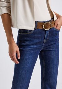 Personne en jeans bleu foncé avec une ceinture en suede marron à boucle ronde, portant une veste beige et un haut de couleur claire, main détendue le long du corps.