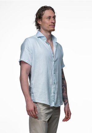 Jonge man met kort krullend haar draagt een lichtblauw overhemd met korte mouwen en een beige broek, met een rozentatoeage op zijn linker onderarm.