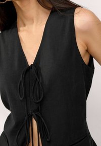 Top in lino nero con scollatura a V profonda e lacci laterali. Presenta una vestibilità comoda e una texture liscia, esaltando la caduta del tessuto.