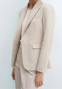 Blazer beige ajusté en tissu lisse, doté d'une fermeture à un bouton, de revers crantés et d'une poche avant, associé à un pantalon assorti.