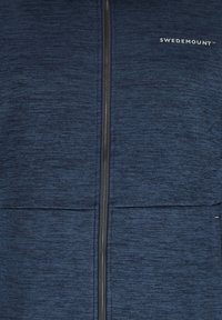 Lähikuva laivastonsinisestä teksturoidusta kangastakista, jossa keskitetty vetoketju ja pieni valkoinen "Swedemount"-logo oikealla rinnassa.