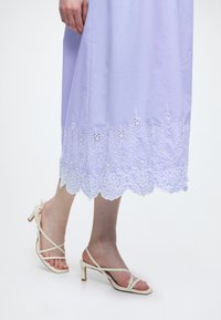 Άτομο που φοράει ανοιχτό μωβ φόρεμα με λουλουδένια δαντέλα στο τελείωμα και μπεζ ψηλοτάκουνα σανδάλια με λουράκια σε λευκό φόντο.