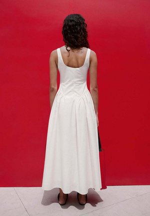 Frau mit lockigem Haar, die ein ärmelloses weißes Kleid und schwarze Absätze trägt, steht mit dem Gesicht zu einer einfarbigen roten Wand und hält eine schwarze Handtasche.