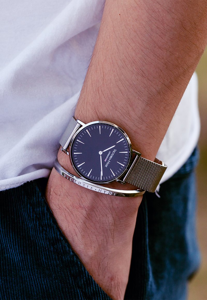 Montre à bracelet en maille argentée avec un cadran rond bleu foncé affichant des index et des aiguilles argentés ; portée avec un bracelet métallique étroit.