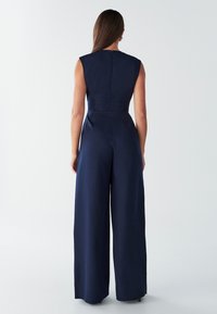 Temno modra kombineza z oprijetim zgornjim delom, širokimi hlačnicami in majhnimi rokavi do ramen. Gladek tekstura; zadrga na hrbtni strani je vidna.