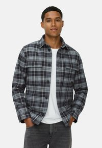 Chemise en flanelle à carreaux gris et noir avec un devant à boutons, deux poches poitrine et un col. Portée par-dessus un t-shirt blanc.