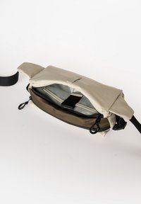 Borsa in tessuto beige con pattina pieghevole, dotata di scomparti con zipper neri e tracolla regolabile. Interno foderato in grigio chiaro.