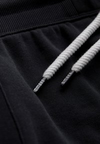 Tissu noir avec cordons tressés blancs, embouts en plastique noir inscrits "OMBRE" et une ceinture élastique.