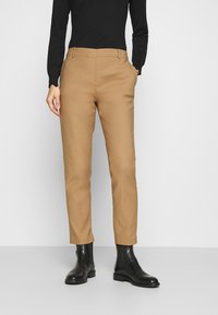 Un pantalon beige slim en tissu lisse avec un devant plat, des poches latérales et des passants de ceinture, associé à des bottines noires.