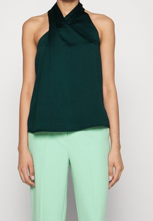 Femme portant un haut dos nu vert foncé et un pantalon taille haute vert clair, debout devant un fond uni.