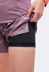 Personne soulève un short lavande pour révéler un short de sport noir texturé avec un petit logo tête de chat noir sur le tissu lavande.