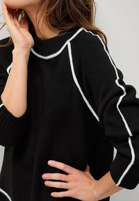 Maglione nero lavorato a maglia con dettagli bianchi lungo le cuciture. Presenta un colletto rotondo e maniche lunghe con polsini a coste. Texture morbida.