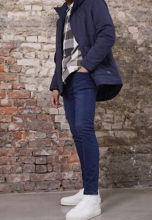 Person trägt navyblaue Jacke, schwarz-weiß kariertes Hemd, blaue Jeans und weiße High-Top-Sneaker und steht vor einer strukturierten Backsteinwand.