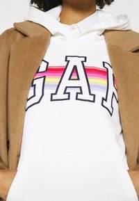 Felpa bianca con grande logo "GAP" in blu e righe arcobaleno, indossata sotto un cappotto beige. Presenta una tasca a marsupio e cappuccio con coulisse.