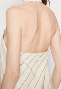 Šaty bez ramínek s krémovou tkaninou a hnědými svislými pruhy. Zadní strana je otevřená, což zvýrazňuje hladkou pleť a minimalistický design.