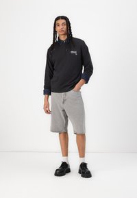 Felpa nera con colletto in denim, pantaloni corti in denim grigio chiaro e scarpe nere chunky; presenta un logo sul petto. Texture del tessuto lisce.