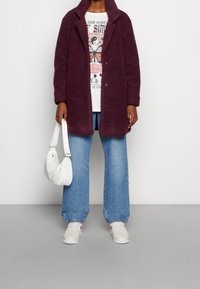 Bordeaux, fluffy jakke med krave, båret over en grafisk t-shirt. Kombineret med brede blå jeans og hvide sneakers, mens der holds en lille hvid taske.