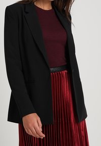 Black tailored blazer featuring a notched lapel, single button closure, and side pockets, paired with a burgundy top and pleated red skirt.