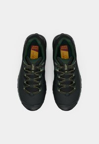 Pantofi sport negri cu talpă din cauciuc texturat, accente verzi și șireturi metalice aurii. Interiorul căptușit și capacul de vârf întărit sunt vizibile.