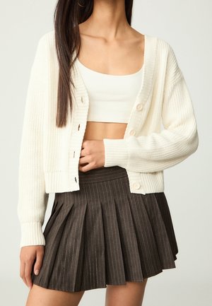 Femme portant un cardigan court blanc, un crop top blanc et une jupe plissée foncée avec de fines rayures, debout devant un fond uni.