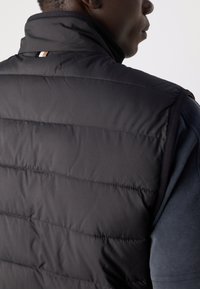 Black puffer vest with a high collar, featuring horizontal quilting, smooth texture, and a small accent label at the back.