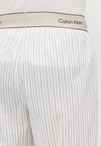 Witte gestreepte boxershorts met een beige elastische tailleband waarop het zwarte "Calvin Klein" logo staat. De stof lijkt lichtgewicht en soepel te zijn.