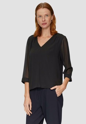 Blusa de gasa negra con escote en V, mangas tres cuartos y puños enrollados. Corte suelto con una textura suave, llevada sobre pantalones azul marino.