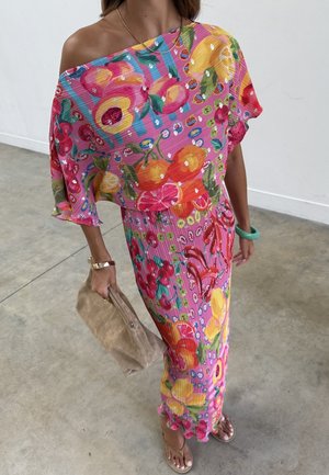 Femme portant une robe colorée à fleurs et épaules dénudées, tenant une pochette beige, avec un bracelet en or et un bracelet vert, debout sur un sol en béton.
