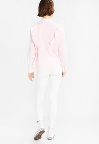 Blusa a righe rosa chiaro con dettagli arricciati sulle spalle, maniche lunghe e colletto, abbinata a pantaloni bianchi aderenti e sneakers.