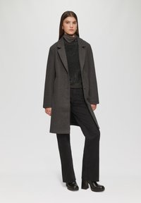Szary wełniany płaszcz z wyrazistymi klapami, zapinany na guziki z przodu, połączony z czarnym teksturowanym swetrem i ciemnymi spodniami. Czarne buty na platformie.
