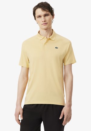 Joven con camiseta tipo polo de manga corta de color amarillo pálido y pantalones negros, de pie frente a un fondo blanco liso.
