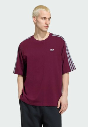 Junger Mann mit kurzen blonden Haaren, der ein dunkelrotes Adidas-T-Shirt mit grauen Streifen an den Ärmeln trägt, mit den Händen in den Taschen steht und nach rechts blickt.