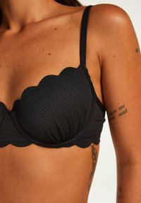 Črn bikini zgornji del s valovitim robom, narejen iz teksturiranega materiala, s prilagodljivimi naramnicami in nabranimi skodelicami. Gladka površina z diskretno heksagonalno vzorčno podrobnostjo.