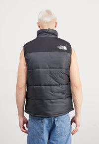 Gilet imbottito nero e grigio con alto collo, caratterizzato da piumini orizzontali e un logo sulla parte superiore sinistra della schiena. Indossato sopra un top senza maniche.