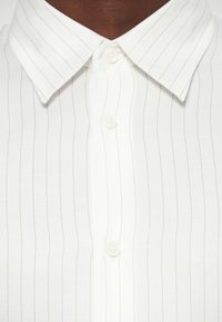 Hvit skjorte med krage, med tynne vertikale striper og to knapper i stangen, laget av glatt stoff.