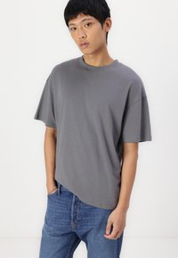 Grå kortärmad t-shirt i bomull med avslappnad passform. Har rund hals och sänkta axlar, tillsammans med blå jeans.
