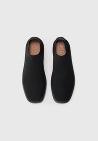 Par med svarte slip-on strikkesko med firkantede tær og flate såler, sett ovenfra på en hvit bakgrunn.
