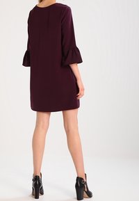 Kvinna klädd i en mörklila knälång klänning med utsvängda trekvartsärmar och svarta högklackade ankelboots, stående med ryggen mot oss.