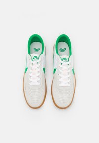 Pár bílých a zelených tenisek Nike SB Heritage Vulc s gumovými podrážkami, viděné z vrchu na bílém pozadí.