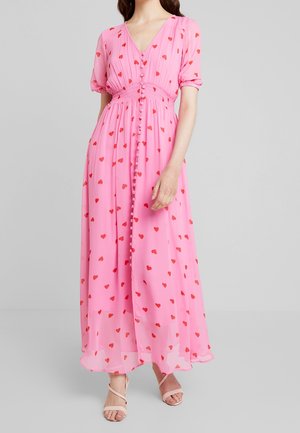 Femme portant une robe maxi rose à motifs de cœurs rouges, avec des boutons sur le devant, des manches bouffantes et des talons beige à brides.