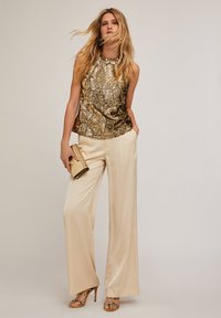 Gouden pailletten mouwloos topje gecombineerd met crèmekleurige broek met wijde pijpen; accessoires omvatten een gouden clutch en beige sandalen met bandjes.