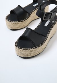 Sandales à plateforme en cuir noir avec semelles en jute tressé, orteils ouverts et brides à la cheville avec grandes boucles, sur fond blanc.