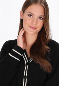 Melna veste ar baltām svītrām, teksturēts adījums, ar V-veida kakla izgriezumu, lielām pogām un kombinēta ar melnām rozēm veidotām auskaru pāri.