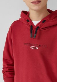 Czerwony hoodie z czarnymi sznurkami, z nadrukiem tekstu, który brzmi "ŚCIEŻKA DŹWIĘKOWA MOJEGO ŻYCIA VOL. 8". Miękki materiał, luźny krój.